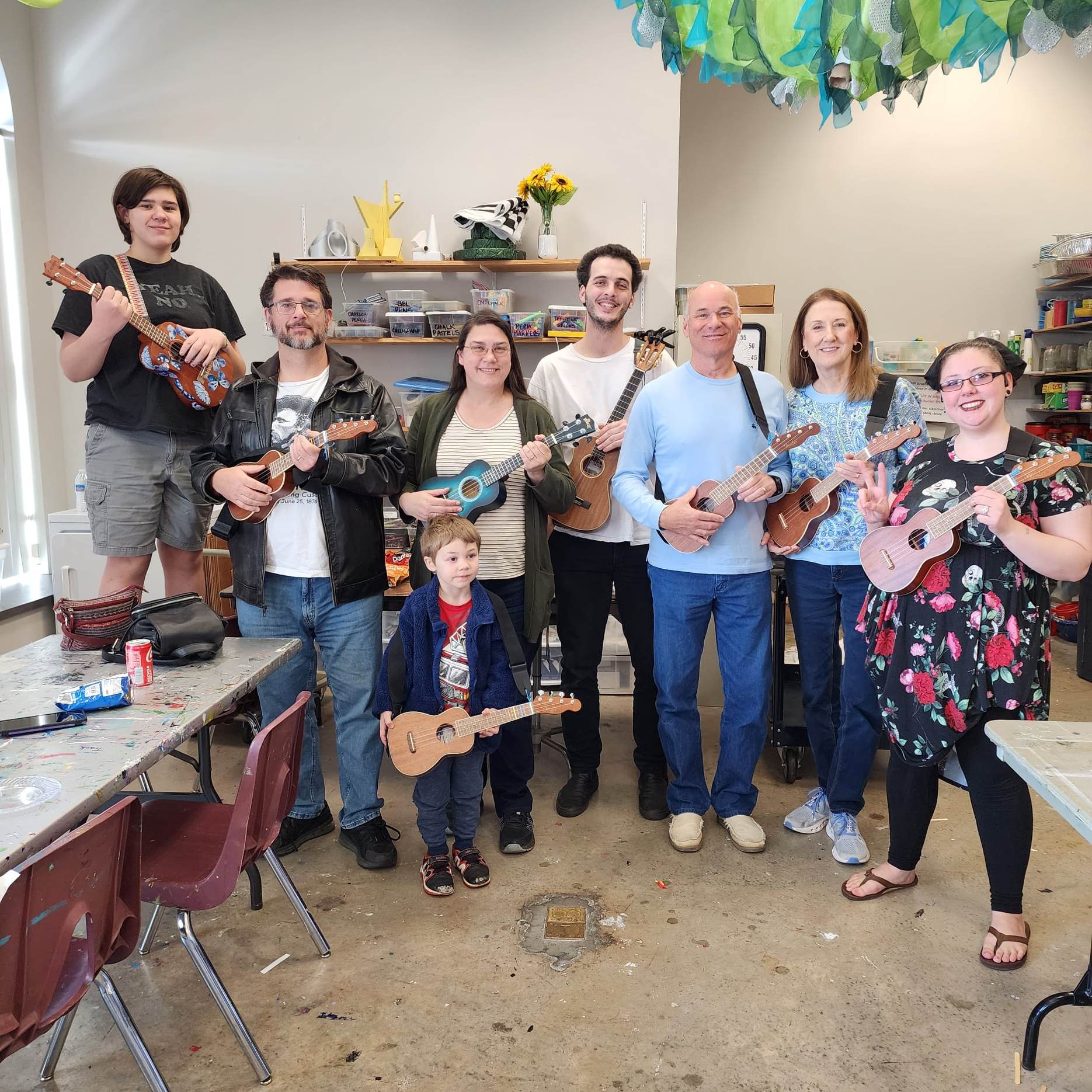 8 people of various ages standing in a row smiling and holding ukuleles