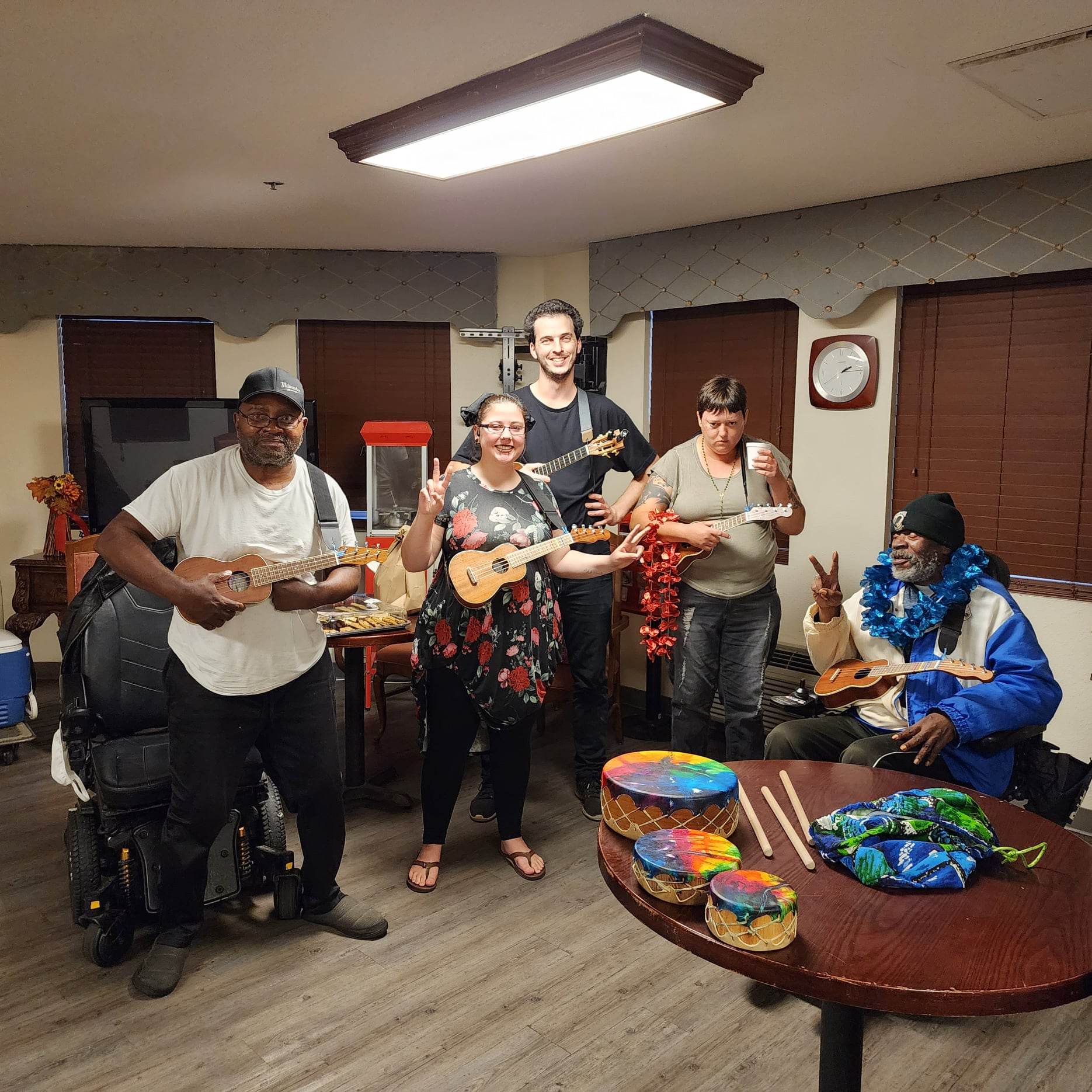 Group of people of various races standing and sitting facing a camera holding ukuleles. The most of the people are smiling. Two of the people are using electric wheel chairs and one of them is standing in front.